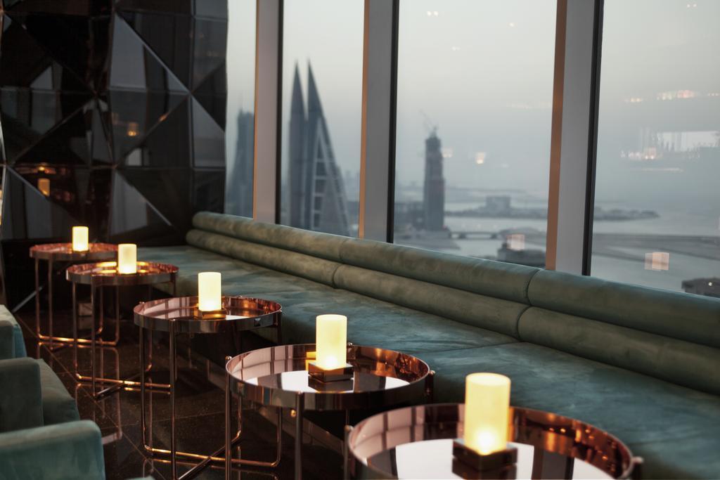 The Domain Bahrain Hotel And Spa - Adults Friendly 16 Years Plus Manama Exterior photo The photo depicts a stylish lounge area with a modern aesthetic. There are several circular tables made of metallic materials, each topped with a small glowing candle. A long, plush seating area in a teal color runs along the window, offering a view 